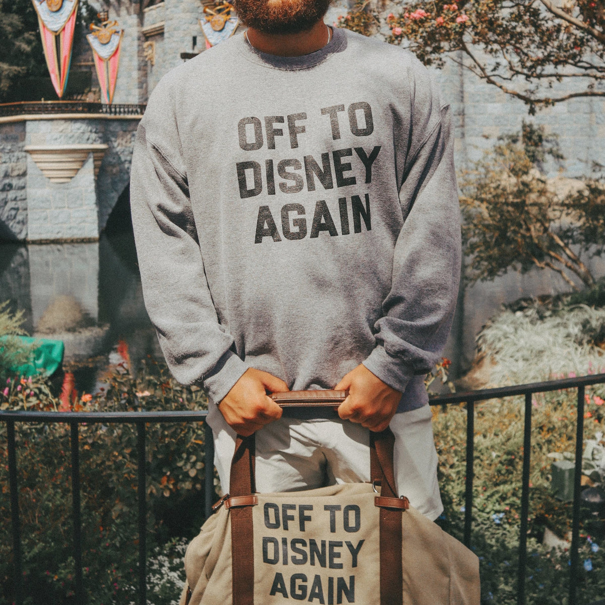 Person wearing a sweatshirt and holding a bag with 'Off to Disney Again' text, standing in front of a castle.