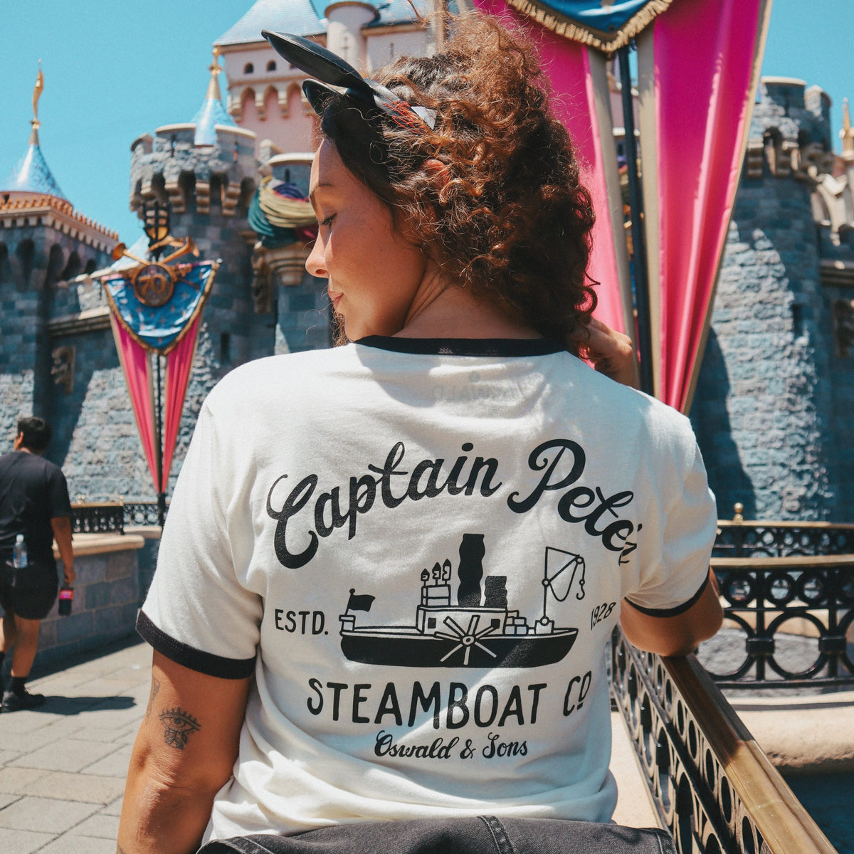 A woman photographed from behind wearing the Steamboat Willie Tee from Oswald & Sons set against a sunny outdoor scene with a castle and colorful banners.