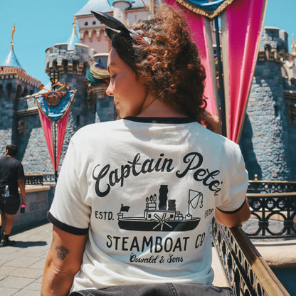 A woman photographed from behind wearing the Steamboat Willie Tee from Oswald & Sons set against a sunny outdoor scene with a castle and colorful banners.