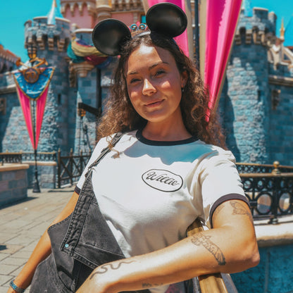 A woman wearing The Steamboat Willie Tee from Oswald & Sons and black overalls, posing in front of a castle with castle-themed flags. She is wearing black Mickey Mouse ears.