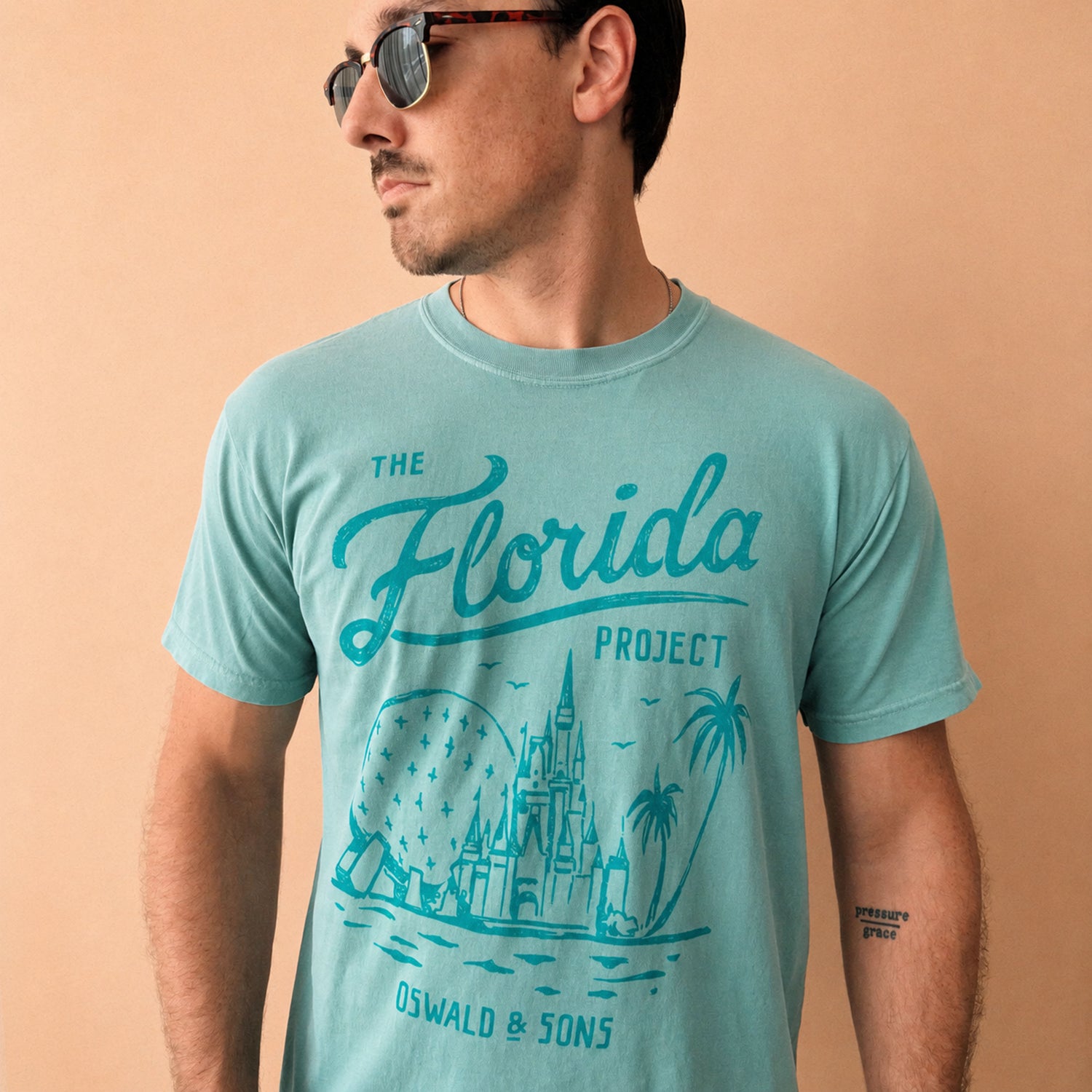A man wearing a seafoam graphic t-shirt with a teal Florida-themed print that reads 'The Florida Project' and 'Oswald & Sons', set against a solid beige background.