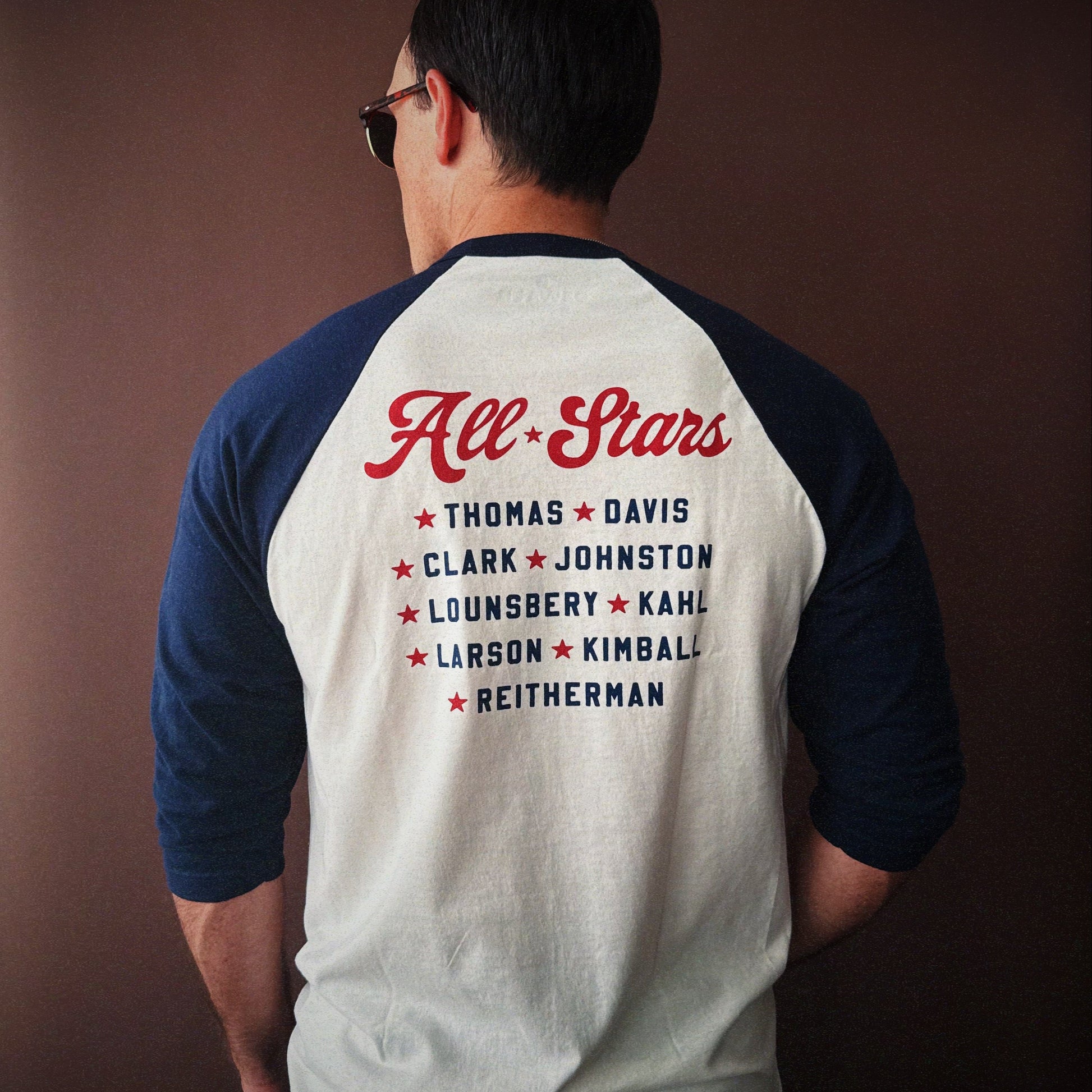Person wearing The Nine Old Men baseball t-shirt with 'All Stars' and names printed on the back against a brown background