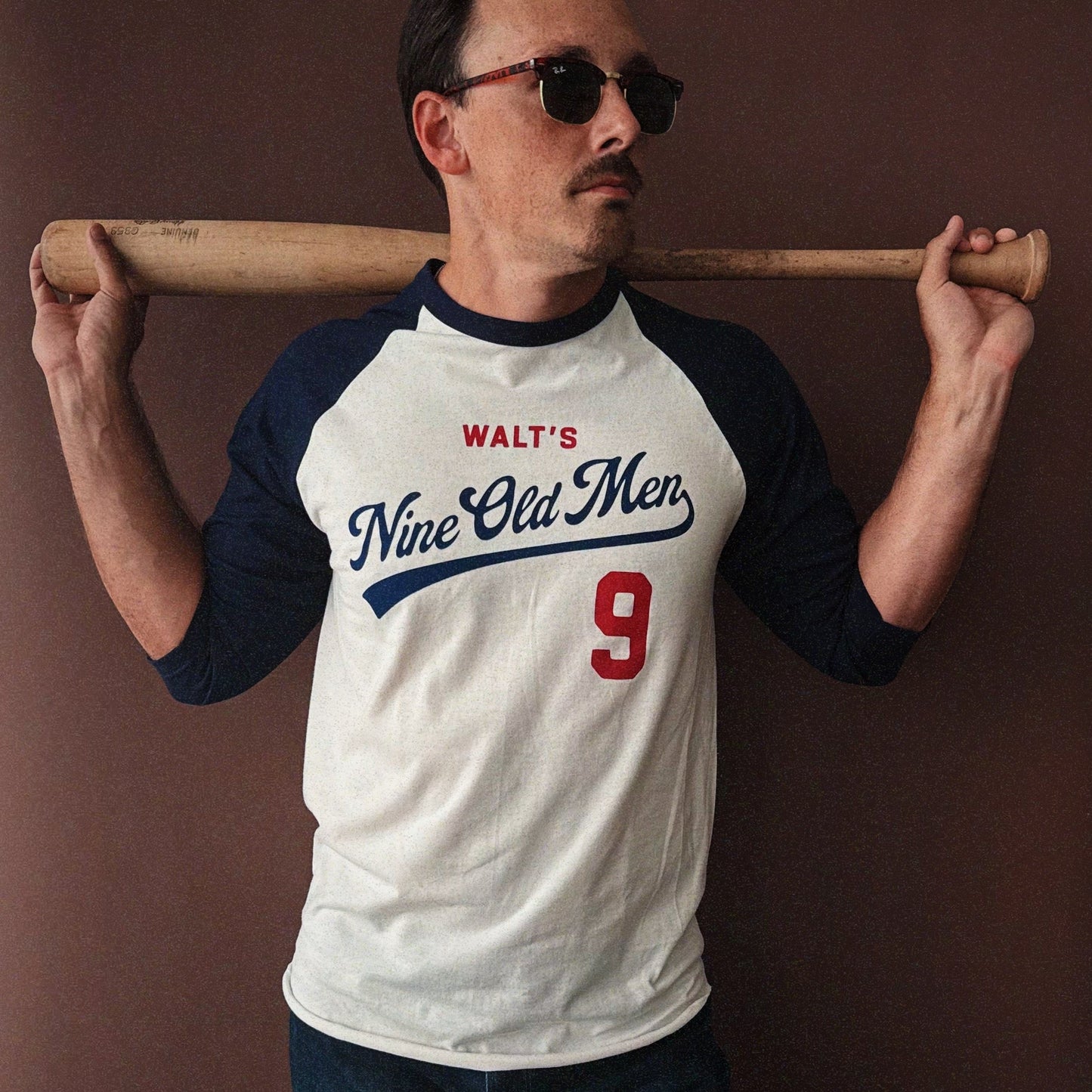 Man wearing the Nine Old Men baseball t-shirt with a bat over his shoulder against a brown background