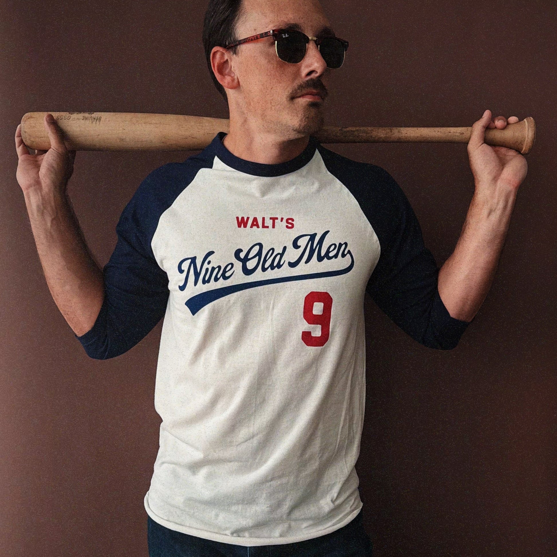 Man wearing the Nine Old Men baseball t-shirt with a bat over his shoulder against a brown background