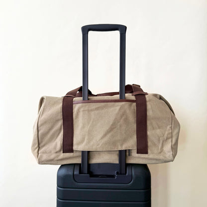 Beige duffel bag with brown straps on top of a black suitcase against a white background